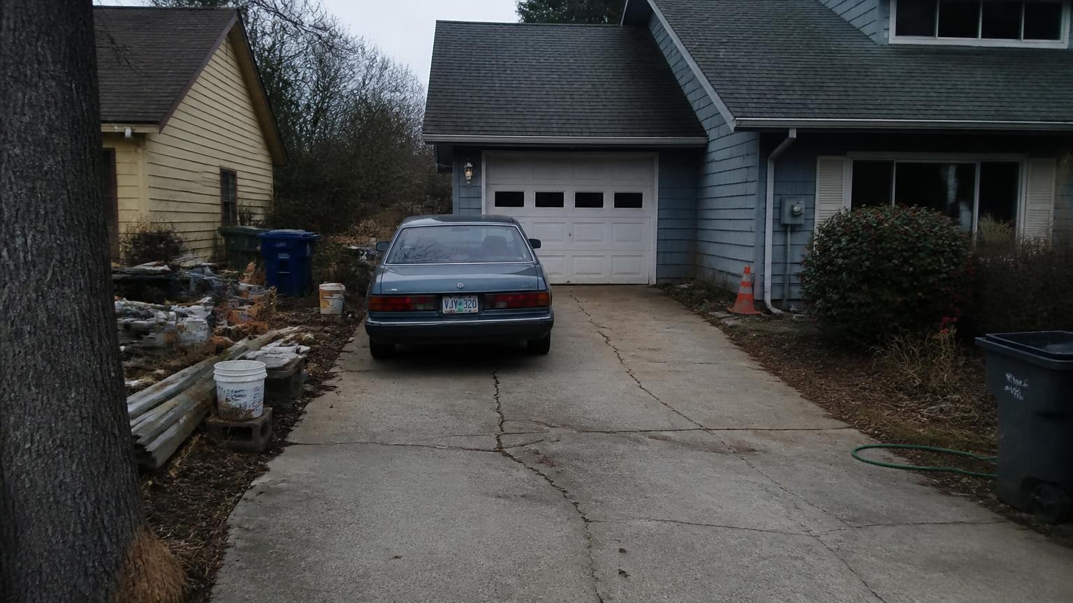 View from the end of a cracked suburban driveway: an aging sedan parked outside a weathered garage, stacked debris and buckets along the side yard.